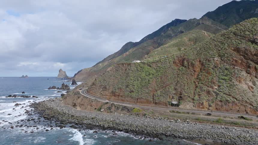 Aerial drone footage captures a scenic highway winding along a rugged mountain coastline as waves crash on the rocky shore