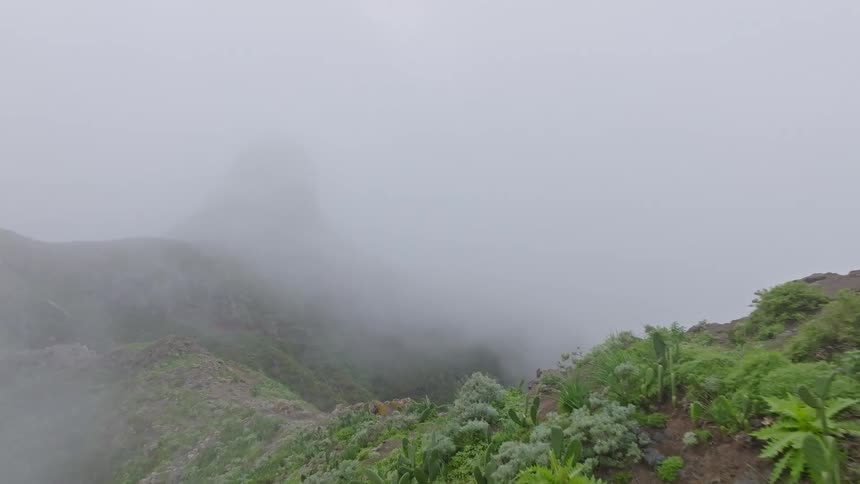 Aerial drone footage flying over a lush green mountainside into a thick, mysterious fog-filled valley