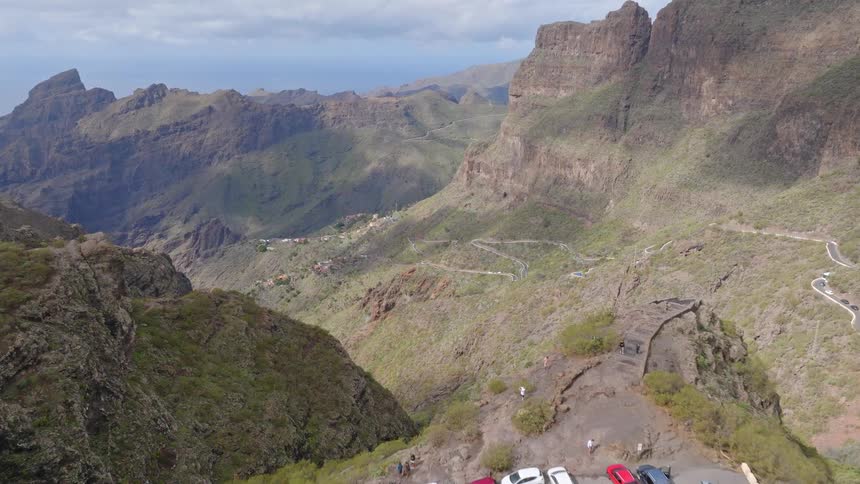 A drone flies forward over a scenic viewpoint with cars, revealing a stunning, rugged mountain canyon with a serpentine road snaking through it
