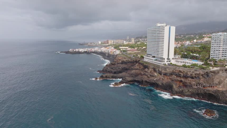 A dramatic aerial view shows modern hotels built along a rugged volcanic coastline under a stormy, overcast sky