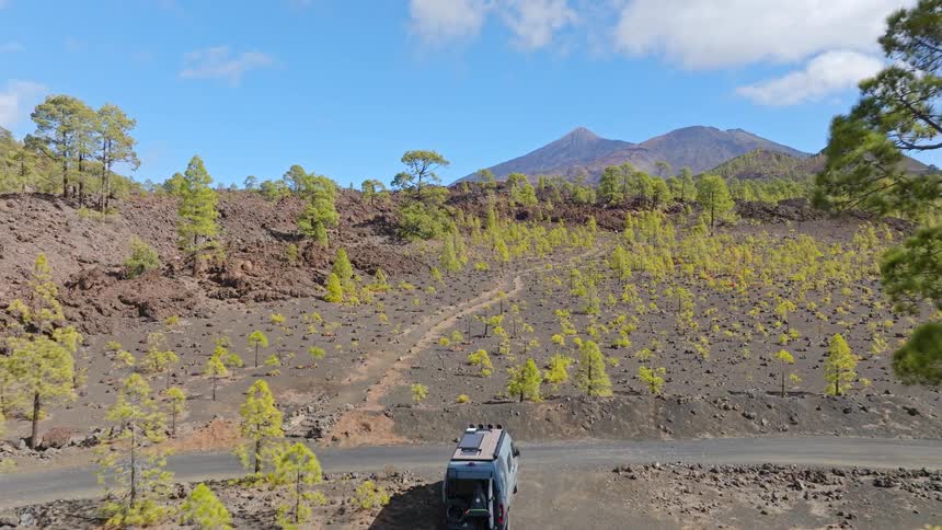 Aerial pull-back reveals a camper van parked on a dirt road amidst a volcanic landscape with vibrant green pine trees and a large mountain