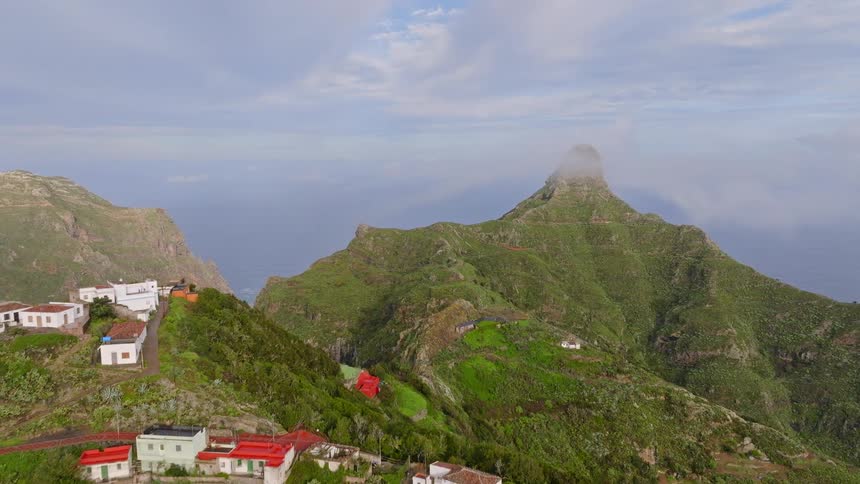 An aerial view pulls back from a mist-covered green mountain peak to reveal a remote village nestled on the cliffs overlooking the vast ocean