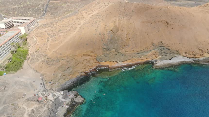 A stunning top-down aerial shot zooms out from swimmers in a turquoise bay to reveal the vast volcanic landscape of Montana Amarilla, Tenerife