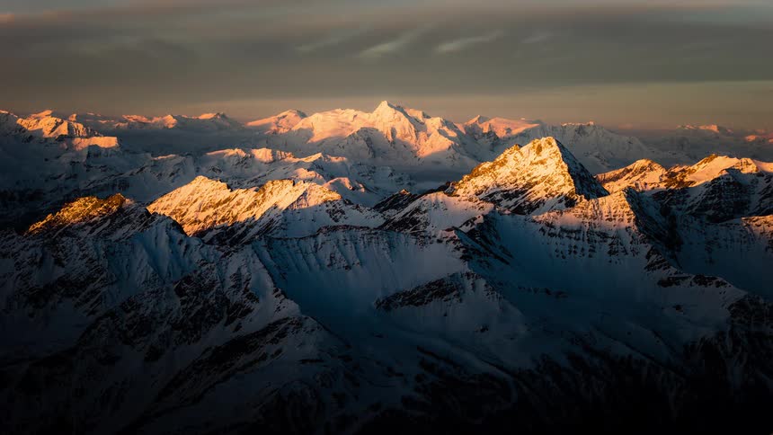 A stunning timelapse shows the golden hour light of sunrise sweeping across a majestic snow-capped mountain range