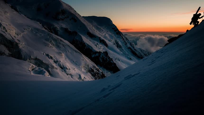 A lone skier carries their skis while climbing a steep, snowy mountain at sunrise, silhouetted against a beautiful orange sky
