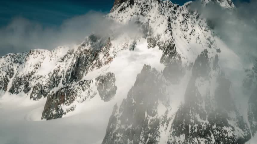 A dramatic tilt-up reveals a majestic, snow-covered mountain peak emerging from swirling clouds against a deep blue sky