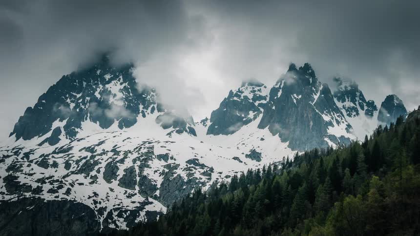 Dramatic time-lapse of dark storm clouds moving over majestic snow-covered mountains and a pine forest