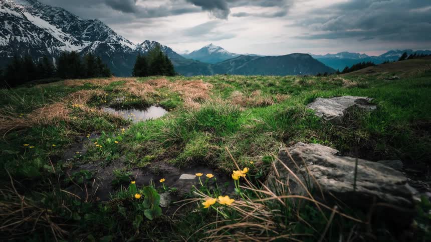 A moody timelapse captures a stream flowing through a green meadow with wildflowers below a majestic snow-capped mountain range under a cloudy sky