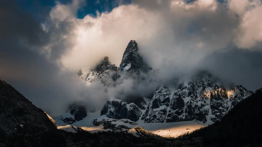 A powerful timelapse shows golden sunlight breaking through dramatic clouds to illuminate a majestic, snow-covered mountain range