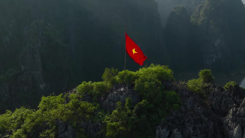 The national flag of Vietnam waves proudly atop a lush, rocky mountain peak in a stunning landscape