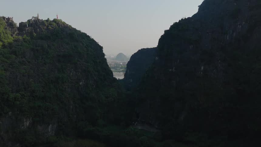 A slow aerial push-in shot through a majestic, shadowed mountain valley reveals a distant river and cityscape in Vietnam