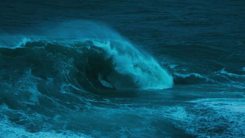 A massive, dark blue ocean wave builds and curls into a powerful break in dramatic slow motion