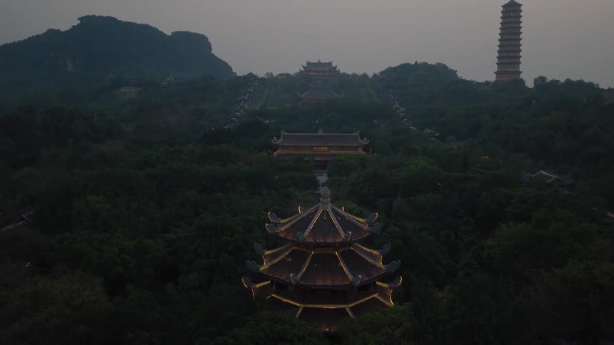 Aerial pullback reveals a vast, illuminated Buddhist temple complex nestled in a misty, mountainous forest at twilight