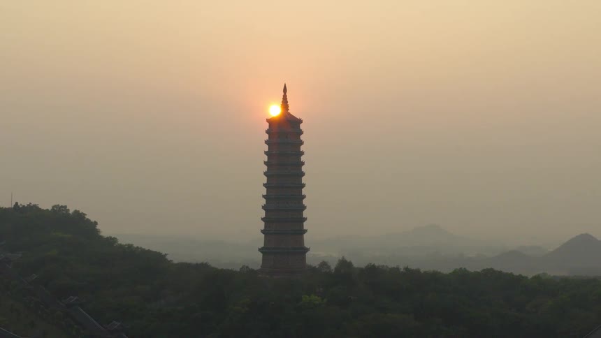 An aerial view reveals the sun rising directly behind a majestic pagoda on a hazy morning