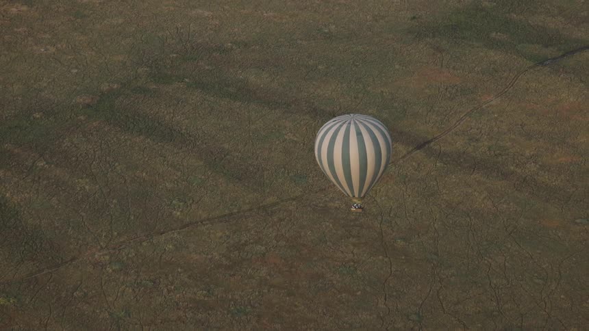 An aerial view shows a striped hot air balloon peacefully floating over the vast, patterned landscape of the African savanna