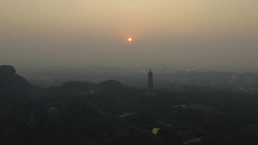A serene aerial view ascends over a vast, hazy Asian landscape, revealing a tall pagoda silhouetted against the setting sun