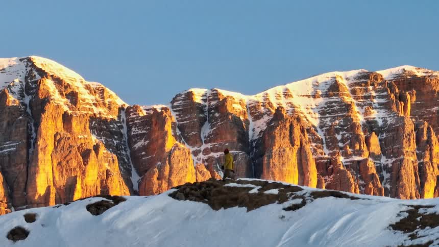 An adventurer in a yellow jacket hikes along a snowy ridge, revealed to be against a backdrop of magnificent sunlit mountains