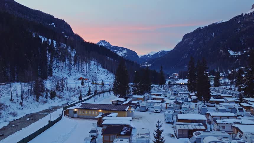An aerial view captures a scenic snow-covered RV campground nestled in a mountain valley during a beautiful winter sunset
