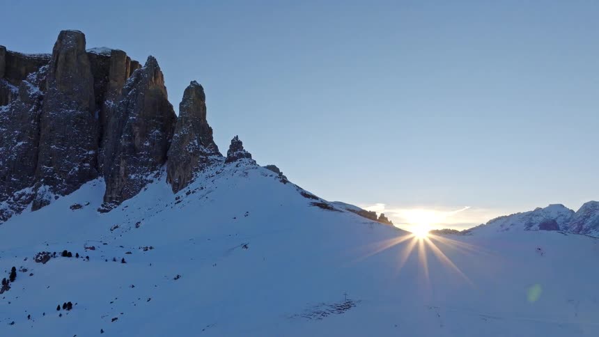 A timelapse shows the sun rising from behind a snow-covered mountain with jagged rock formations against a clear blue sky
