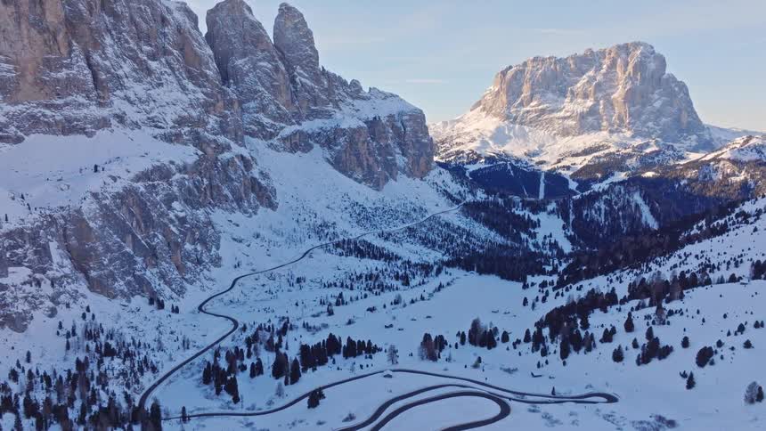 An aerial view reveals a winding road through a majestic, snow-covered mountain pass in the Alps during winter