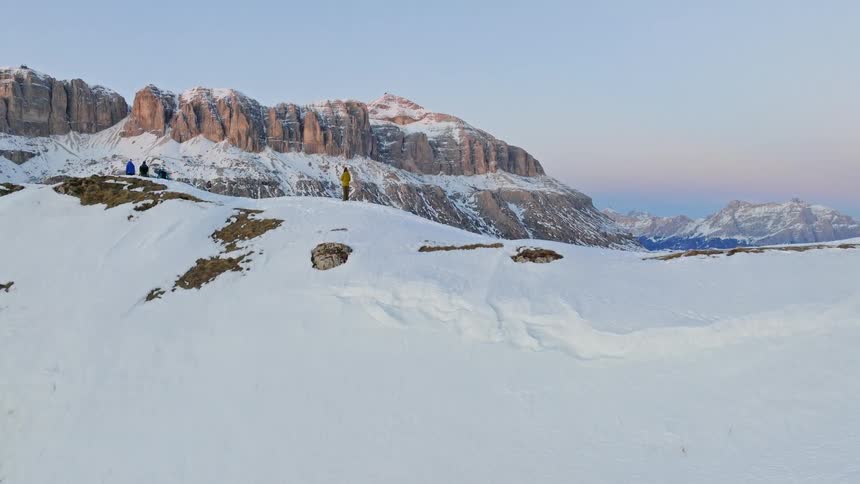 An epic aerial reveal shows hikers standing on a snowy ridge, dwarfed by the majestic Dolomite mountains at sunrise
