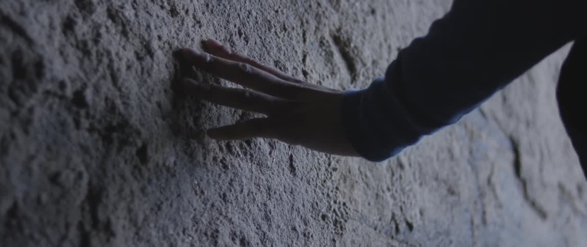 A woman tracing her hand against a wall that crumbles as she touches it