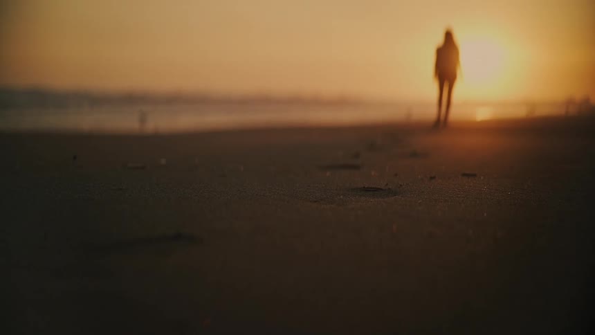 An out-of-focus person stands on a tranquil beach during a golden sunset, with footprints in the sand leading towards them
