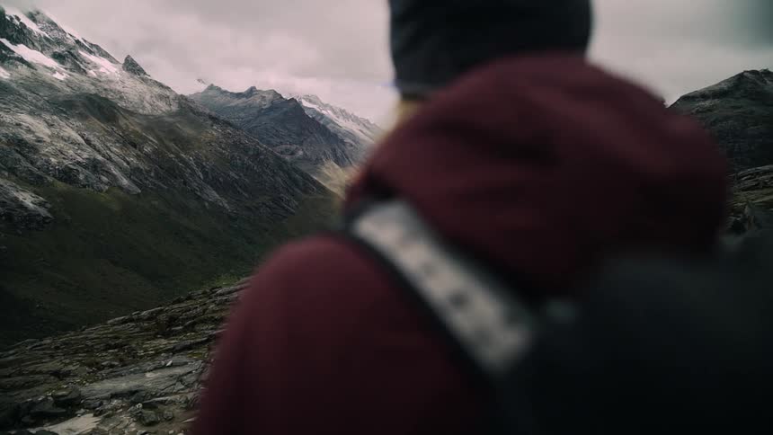 A slow motion rack focus shot from behind a hiker reveals a breathtaking view of a snow-capped mountain range and a valley with a turquoise lake