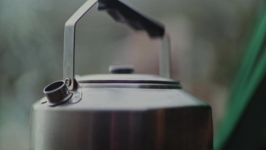 A stainless steel tea kettle boils, releasing gentle steam from its spout in a rustic outdoor setting