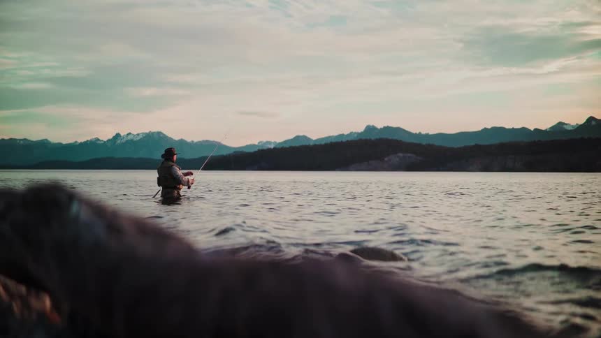 A lone fly fisherman casts his line while wading in a serene lake against a backdrop of mountains at dusk