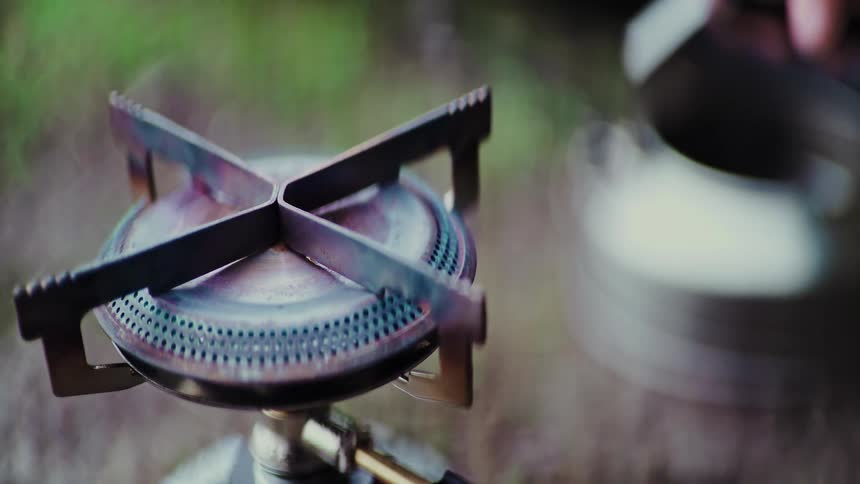 Close up of a person lighting a portable camping stove and placing a metal kettle on the flame in slow motion