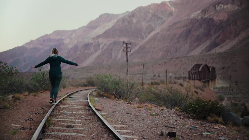 A woman carefully balances on an abandoned railway track curving through a vast and remote mountain landscape