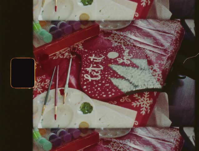 A nostalgic, top-down view of a Christmas arts and crafts project with paints and brushes on a festive tablecloth