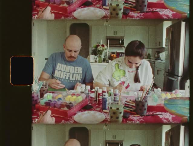 A couple sits at a festively decorated table painting Christmas ornaments together in this vintage-style footage