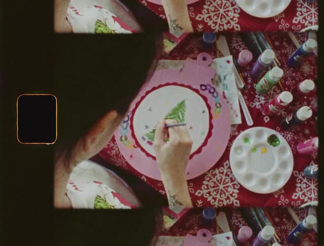 An overhead view with a vintage film aesthetic shows a person painting a Christmas tree on a pink holiday ornament