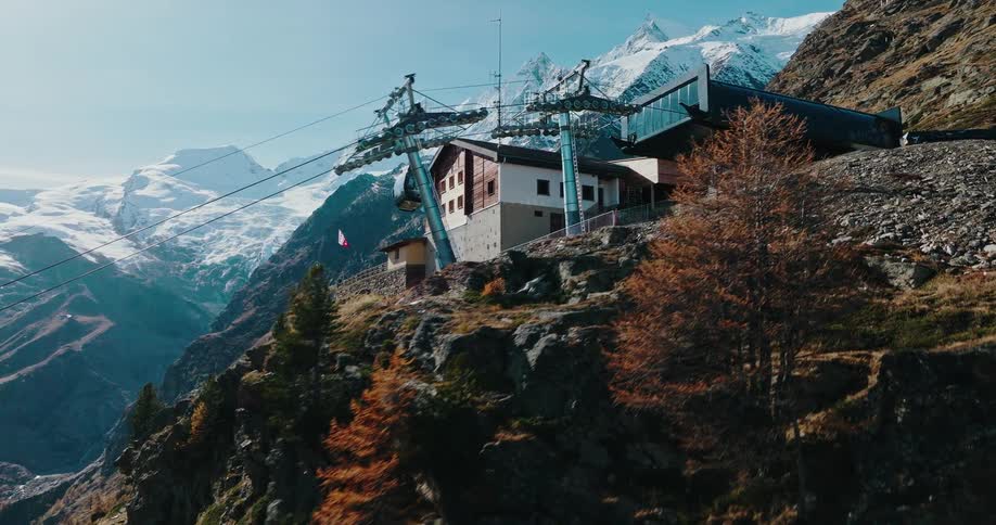 An aerial shot follows a modern gondola arriving at a high-altitude station with majestic snow-capped mountains in the background
