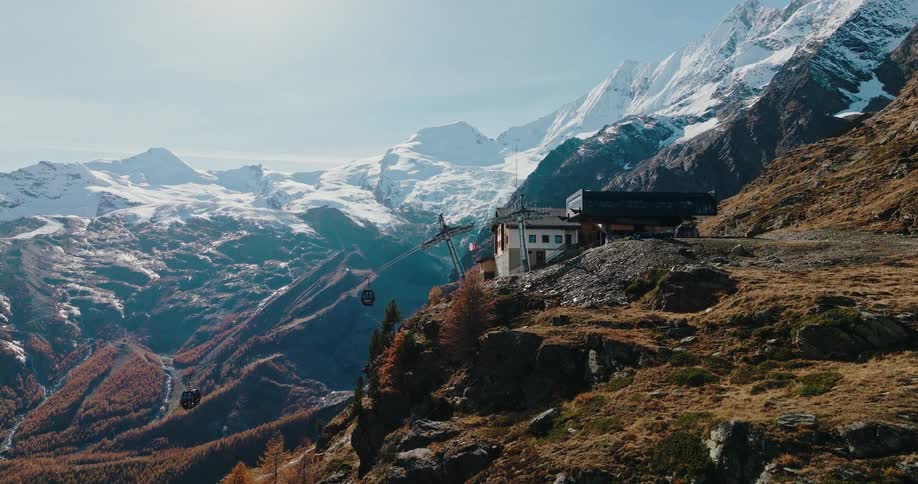 An aerial view shows a gondola arriving at a mountain station, with the majestic, snow-capped Swiss Alps in the background