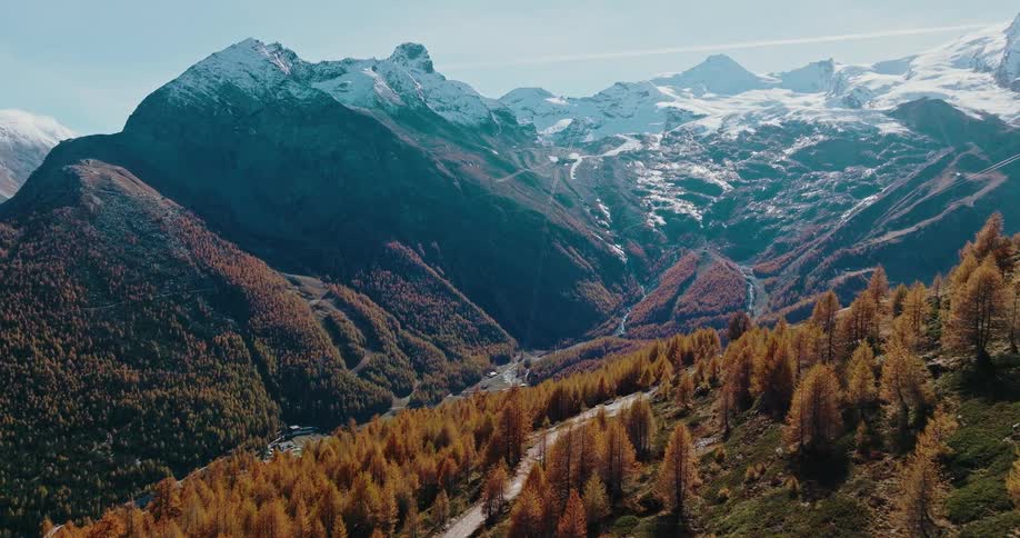 An aerial view reveals a winding road through a golden autumn forest in a valley surrounded by majestic snow-capped mountains