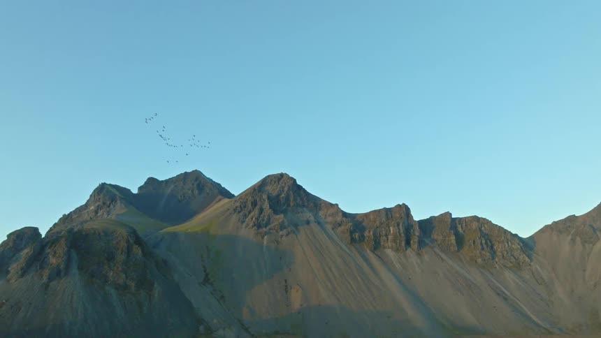 A flock of birds flies past the dramatic Vestrahorn mountain, with an aerial reveal of the stunning Icelandic coastal landscape at sunrise