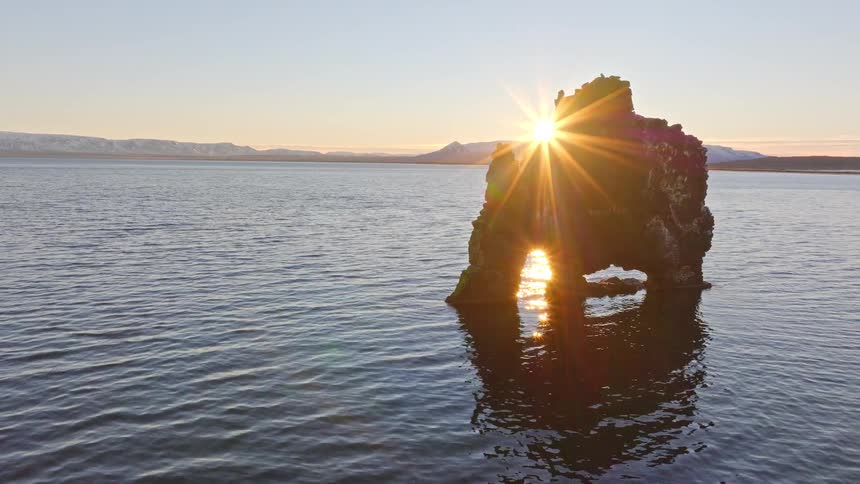 Aerial view of the sun rising and creating a starburst over Iceland's iconic Hvitserkur sea stack, reflecting in the calm ocean below
