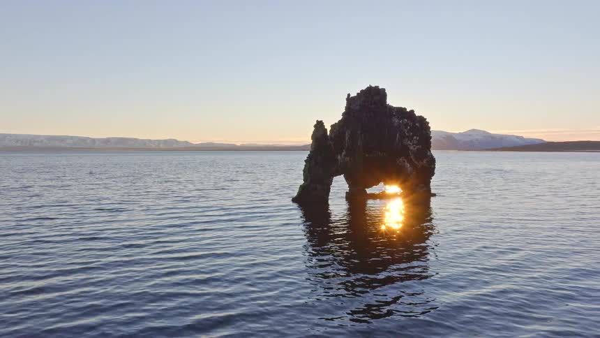 A majestic aerial pullback reveals the iconic Hvitserkur sea stack in Iceland, perfectly framed by the rising sun shining through its arch over the calm ocean
