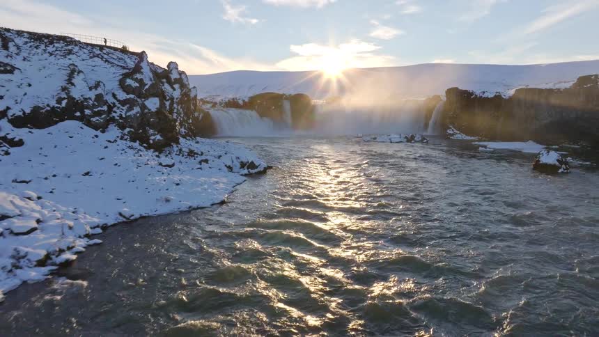 A stunning aerial view reveals a majestic waterfall in Iceland, surrounded by a snow-covered landscape during a beautiful winter sunset