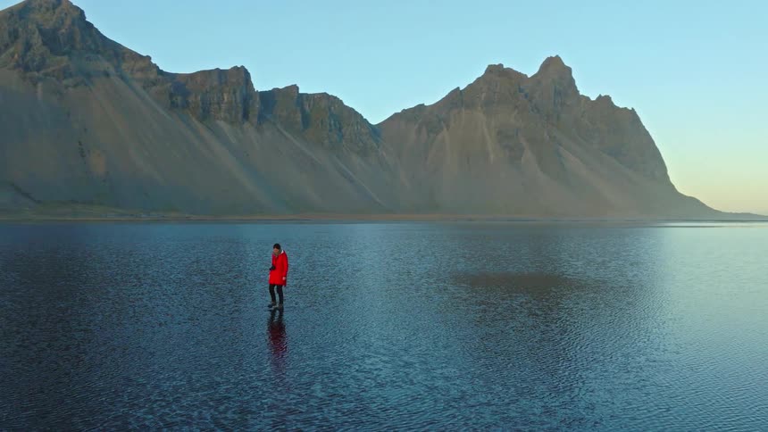 A majestic aerial view captures a lone traveler in a vibrant red coat walking across serene, reflective waters before the stunning Vestrahorn mountain in Iceland