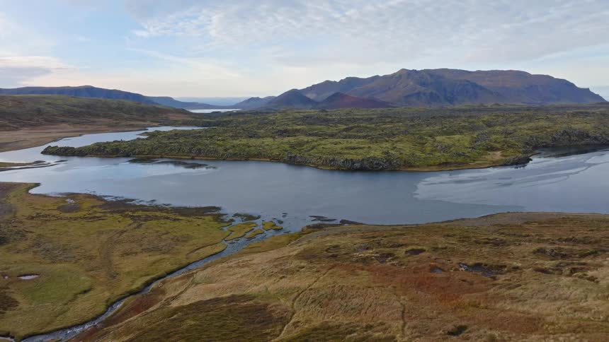 A stunning aerial pullback shot showcases the vast and rugged Icelandic landscape with its serene lakes, mossy lava fields, and mountain ranges