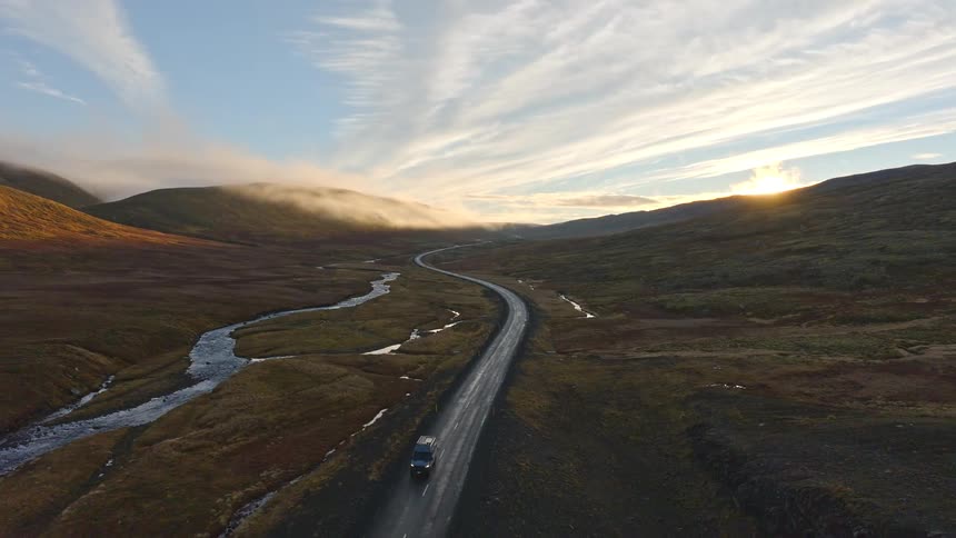 An aerial view follows a lone car driving on a scenic, winding road through a vast mountain valley at sunrise