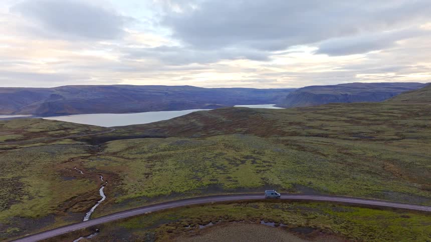 An aerial shot follows a camper van driving along a remote road through a vast, mossy landscape overlooking an Icelandic fjord under an overcast sky