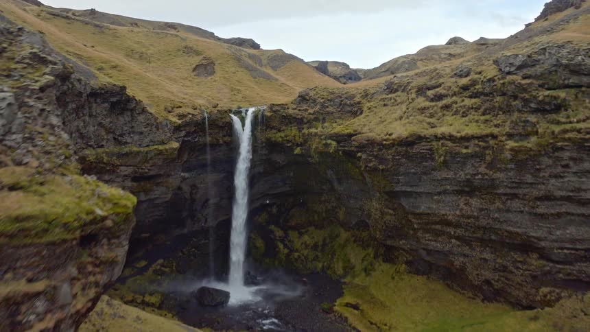 An aerial drone flies past a couple admiring a magnificent waterfall before soaring over the falls and through a beautiful Icelandic canyon