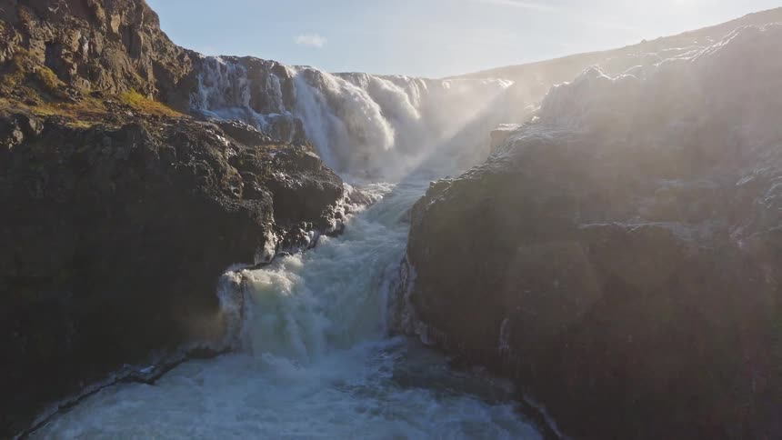 A slow aerial pullback reveals a powerful waterfall cascading through a rugged, sunlit canyon with mist rising into the bright sky