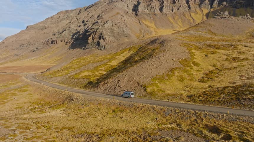 An aerial shot follows a silver camper van as it drives along a scenic dirt road through a vast, mountainous landscape