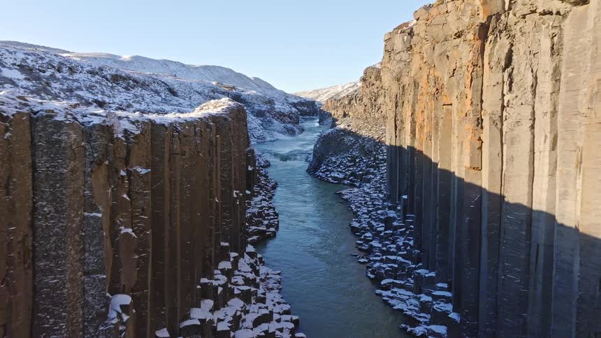 A slow aerial push-in shot reveals the majestic Studlagil Canyon in winter, with its iconic basalt columns covered in snow and a river flowing below